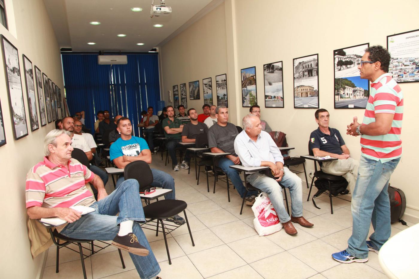 Sala de aula com várias pessoas sentadas ouvindo instrutor que está à frente de todos. #Pracegover