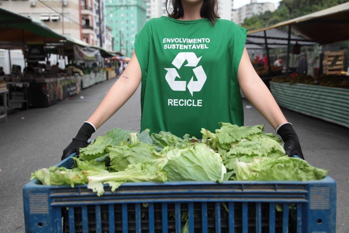 feira livrea ao fundo com destaque para mulher (sem aparecer o rosto) segurando engradado com verduras. Ela usa um colete onde se lê Envolvimento sustentável. Recicle. #paratodosverem 