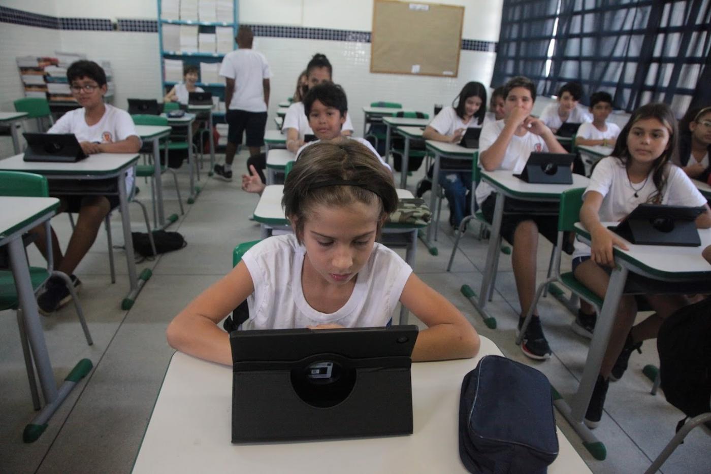 alunos estão sentados nas carteiras diante de tablets abertos. #paratodosverem