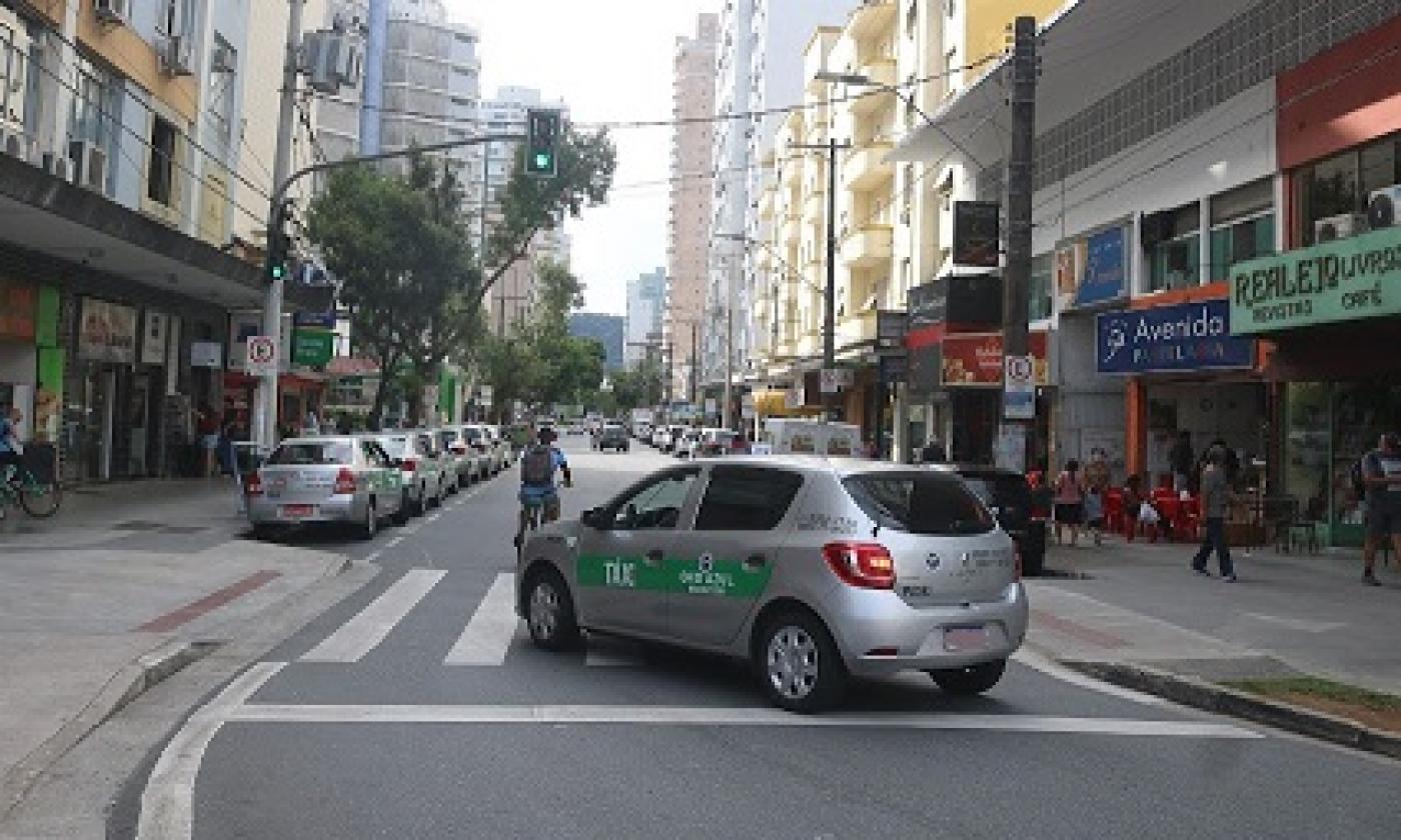 táxi está passando em rua sobre faixa de pedestres. Ao lado, há estabelecimentos comerciais. #paratodosverem