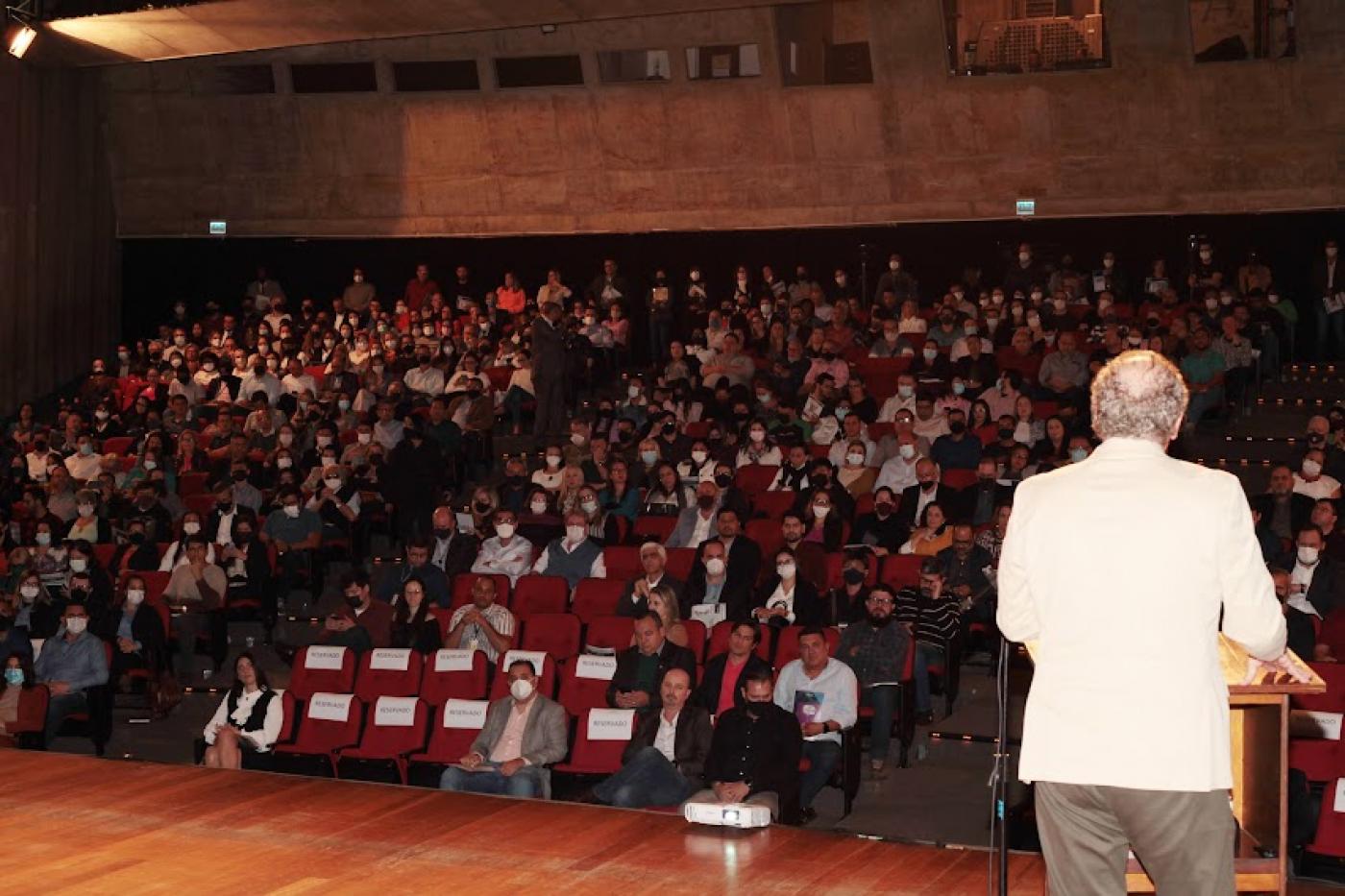 Auditório do Teatro Municipal com lotação quase máxima. No palco, de costas, um homem está à frente de uma tribuna falando ao público. #paratodosverem