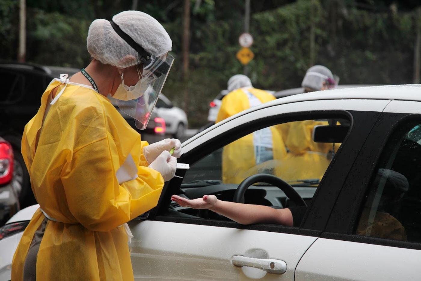 Mulher paramentada com avental, luva, gorro e máscaras faz teste em pessoa dentro de carro. #Paratodosverem