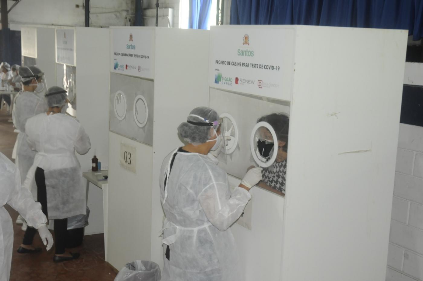 pessoas estão passando por teste nasal de covid atrás de biombos. Técnicos vestidos de branco procedem o exame do lado externo. #paratodosverem