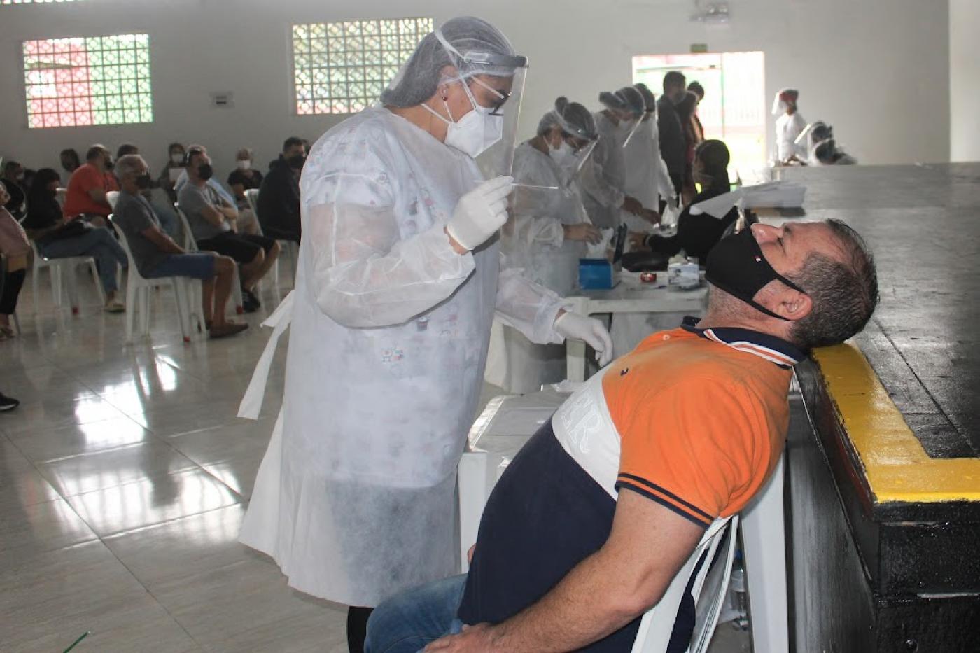 mulher usando avental, máscara, luva e touca prepara para introduzir tipo de cotonete nasal em homem que está sentado. Ao fundo, outras pessoas esperam pelo exame. #paratodosverem