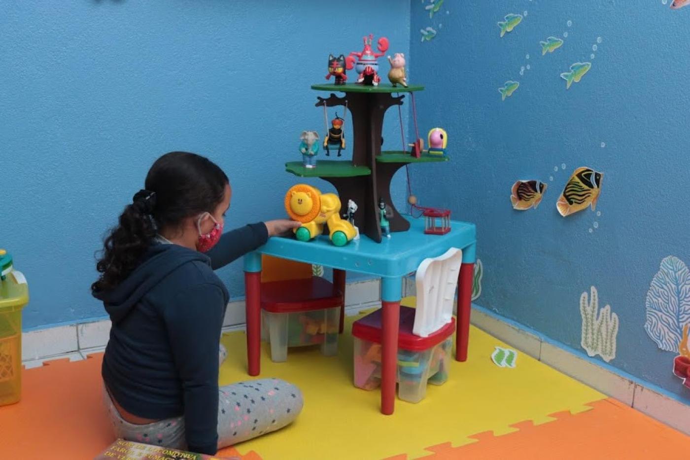 meninas está mexendo em brinquedo infantil em uma sala com piso emborrachado. Ela está de costas para a foto. #paratodosverem