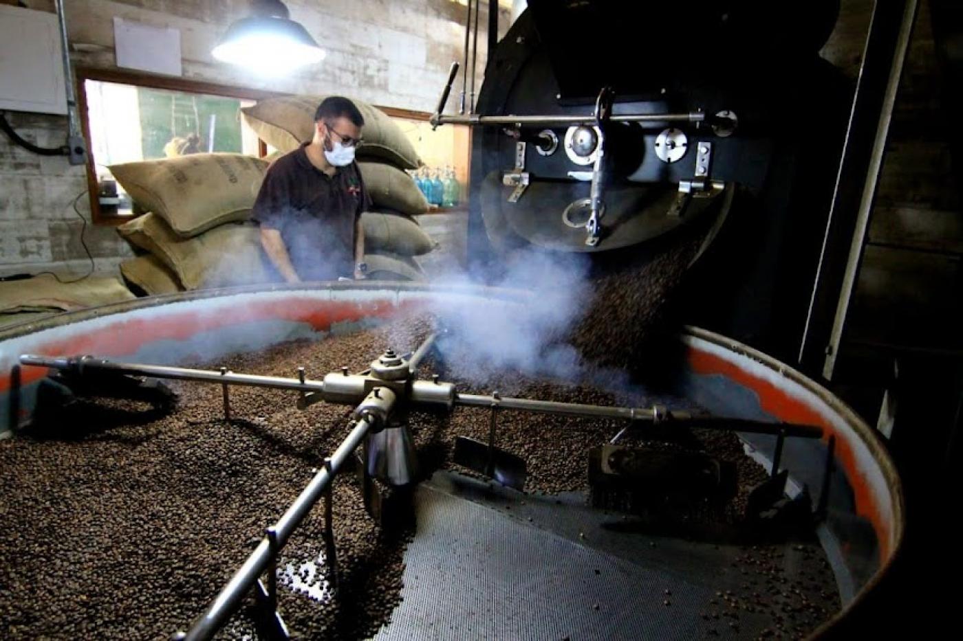 Equipamento de torra de café com diversos grãos. Máquina está em movimento. Homem está ao fundo diante de sacas de café empilhadas. #paratodosverem