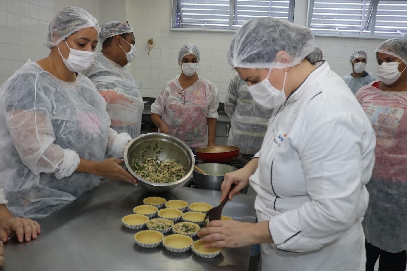 mulheres em primeiro plano coloca recheio em forminhas de torta sobre uma mesa. Outra mulher segura uma cumbuca com recheio. #paratodosverem