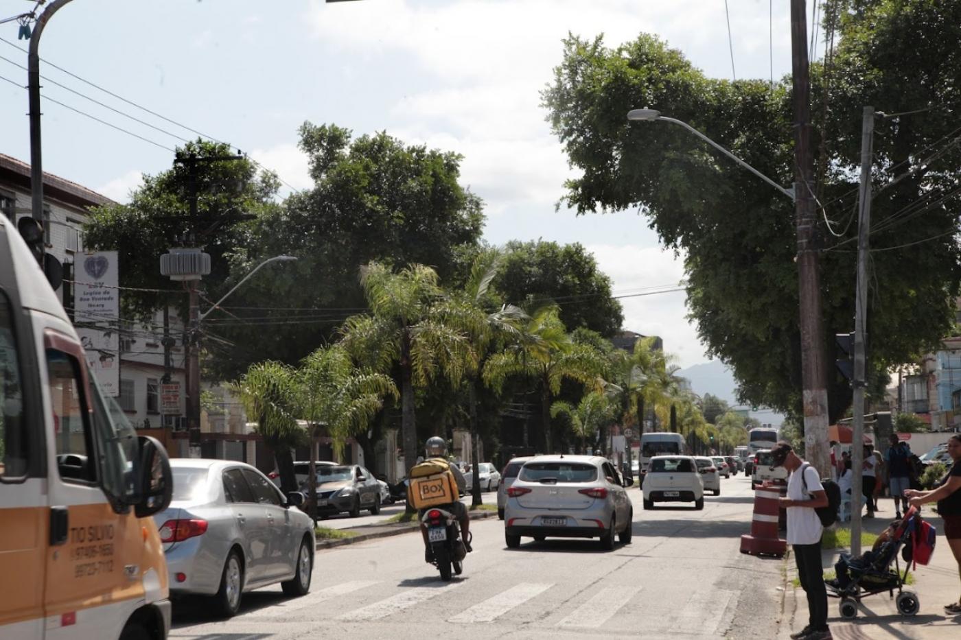 veículos trafegando em pista. Pessoas paradas em frente a uma faixa de pedestres aguardando para a travessia. #paratodosverem