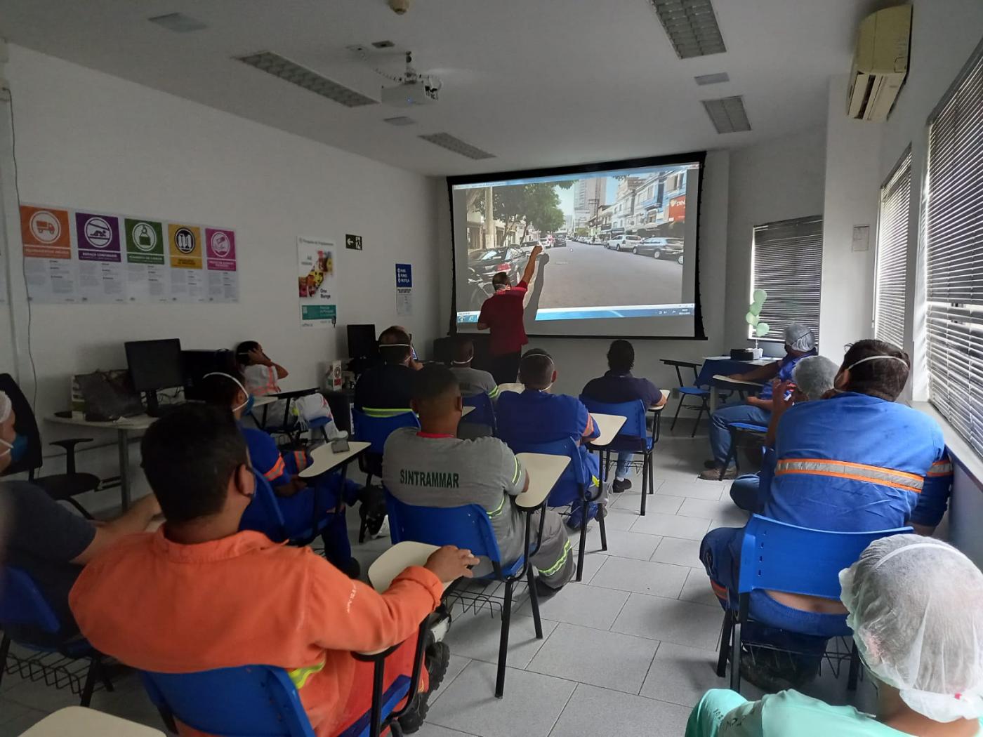 Profissionais sentados em uma sala observam explicações de homem apontando para telão #paratodosverem