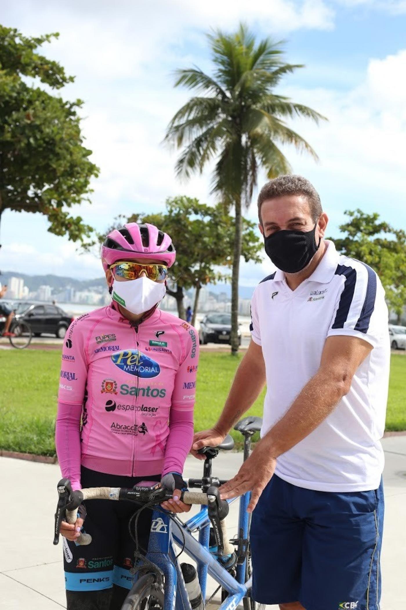 Cláudio está em pé ao lado de bicicleta e de uma jovem atleta. Eles estão no jardim da orla. #paratodosverem