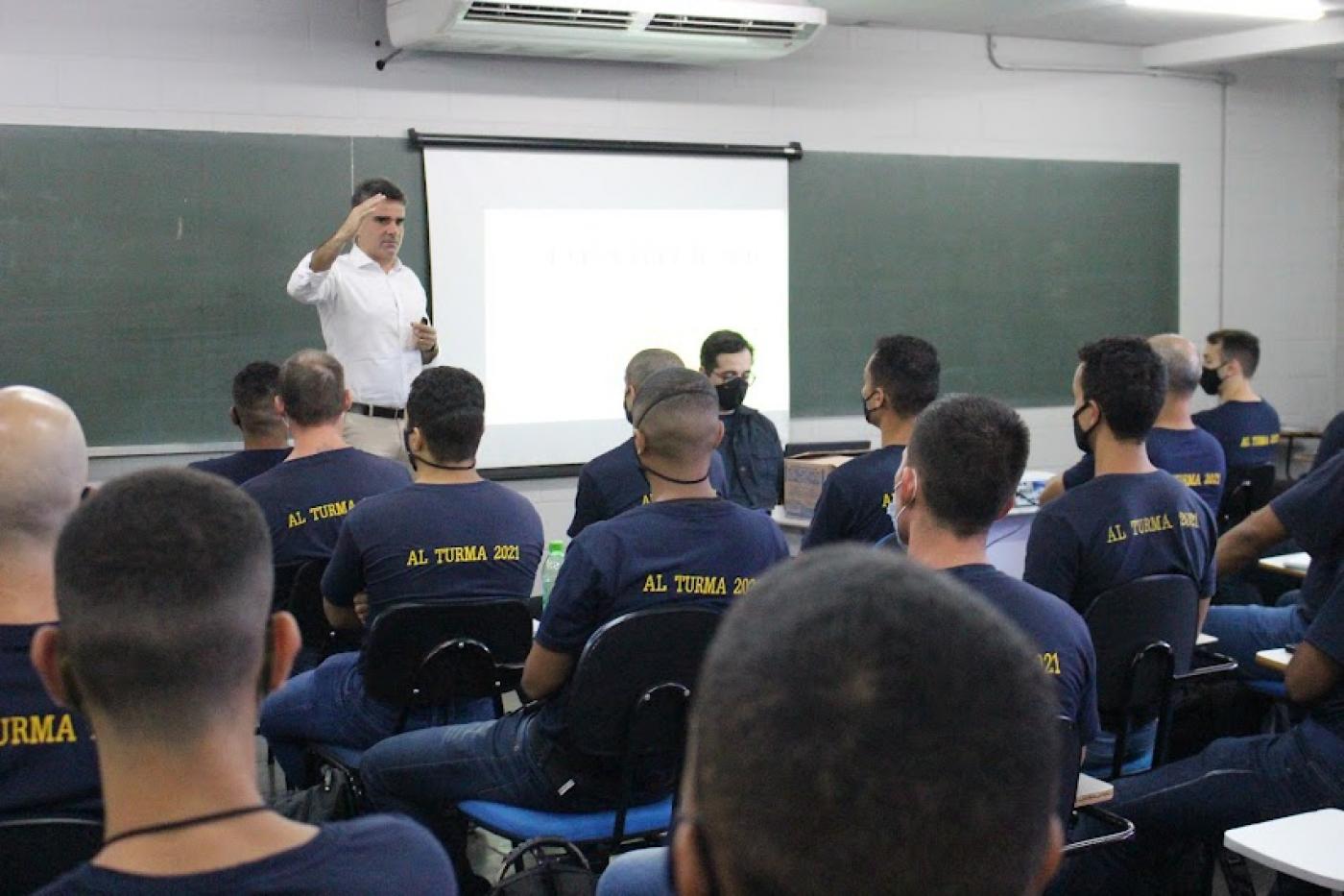 Homem está em pé, ao lado de telão, com a mão direita erguida ao falar com os guardas que estão sentados. #paratodosverem