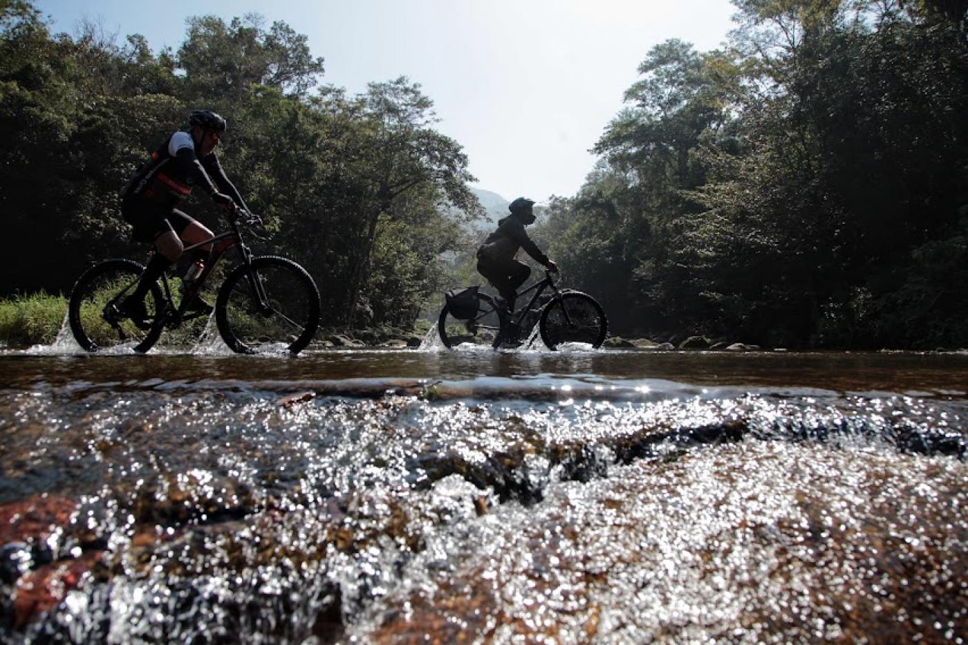 duas bicicletas passando sobre pequeno córrego em pedras com vegetação ao fundo