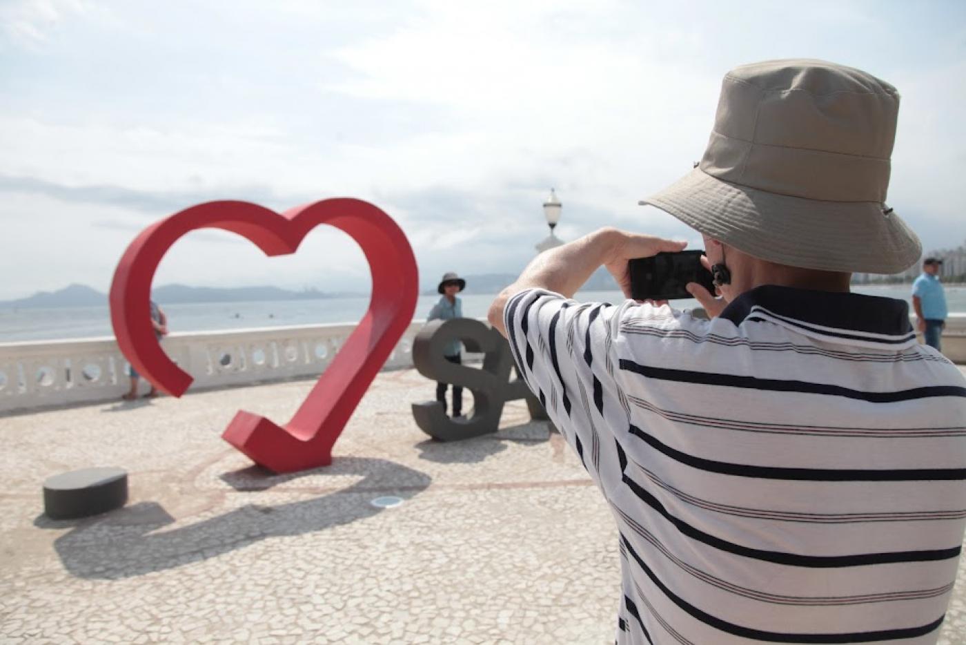 turista usando chapéu (de costas para a foto) usa celular para fotografar pessoa que posa atrás de monumento amo santos, diante da mureta da ponta da praia. #paratodosverem
