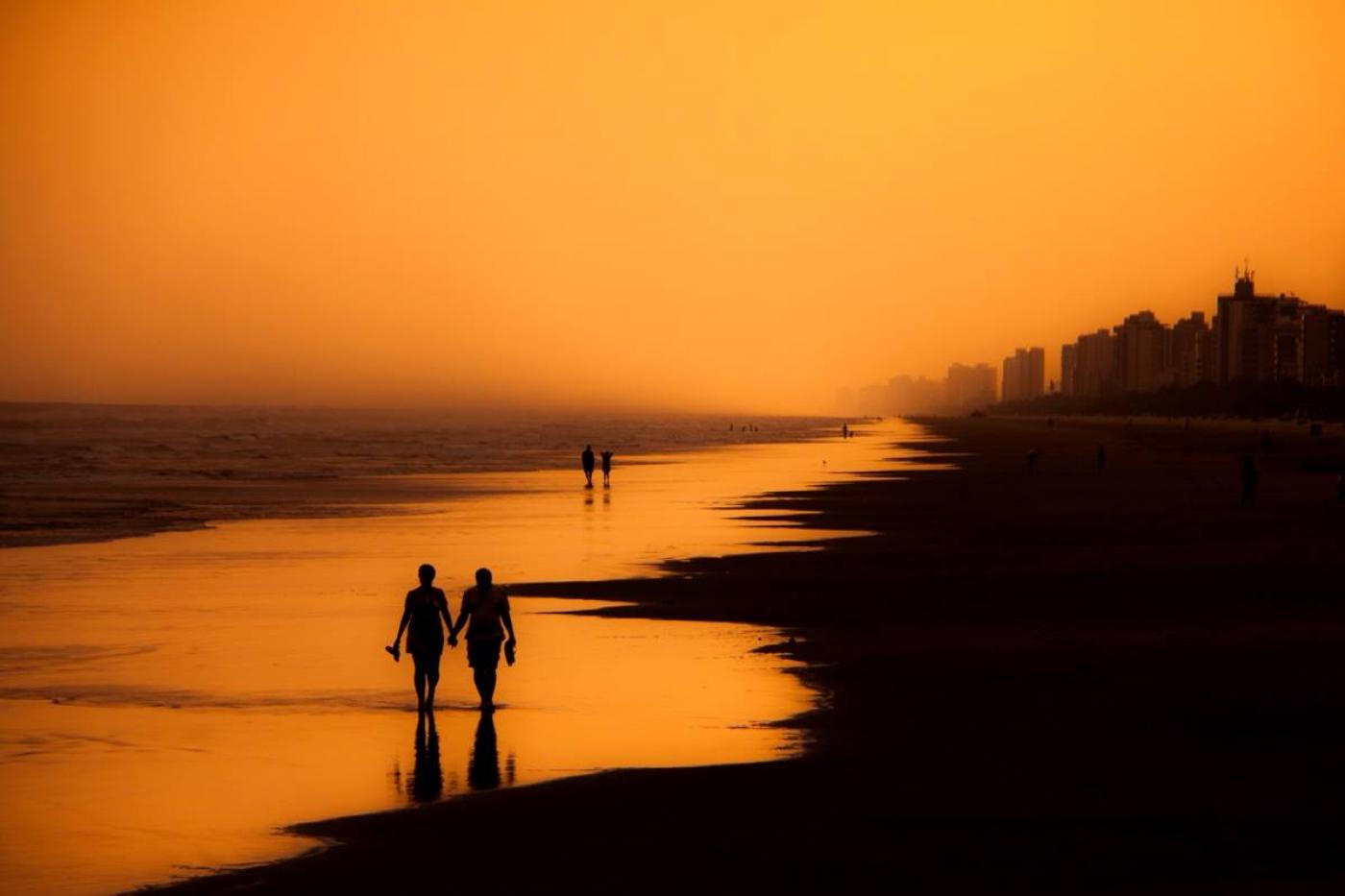 pessoas caminhando à beira-mar. Combinação de corres laranja, do sol, e o escuro da areia se destacam com as silhuetas de duas pessoas caminhando  #paratodosverem