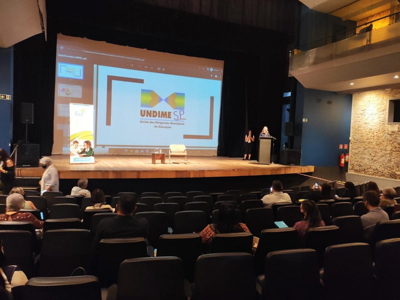 teatro com pessoas sentadas na plateia e duas pessoas no palco. Ao fundo há um telão onde se vê reproduzido o logotipo da Undime 