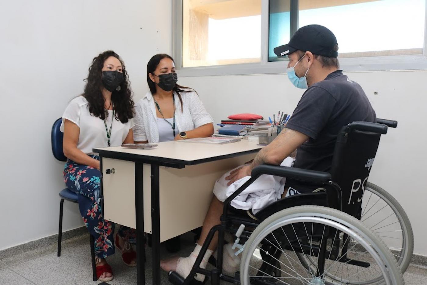 homem está em cadeira de rodas com curativos nos dois pés. Ele está diante de mesa. atrás da mesa estão sentadas duas mulheres. #paratodosverem