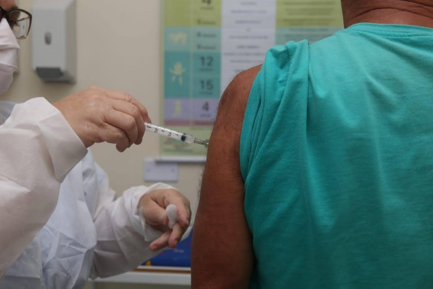 imagem em close foca em mãos de mulher vacinando homem no braço. A manga da camiseta dele está arregaçada.. #paratodosverem