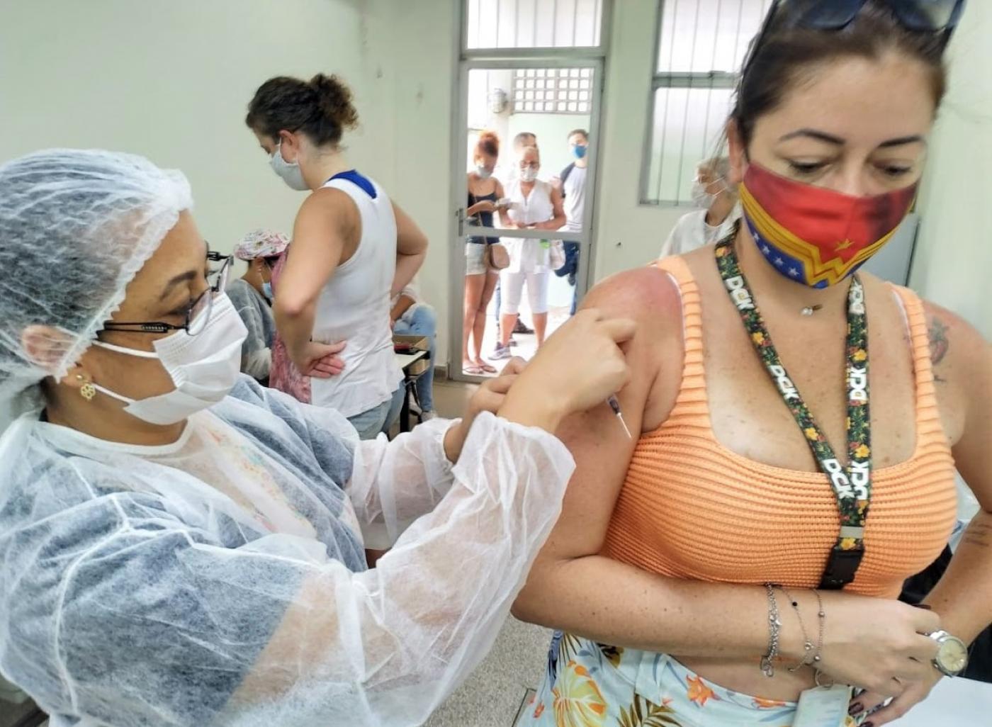 Mulher usando máscara e crachá está sendo vacinada por outra mulher usando avental, máscara e touca. #Paratodosverem