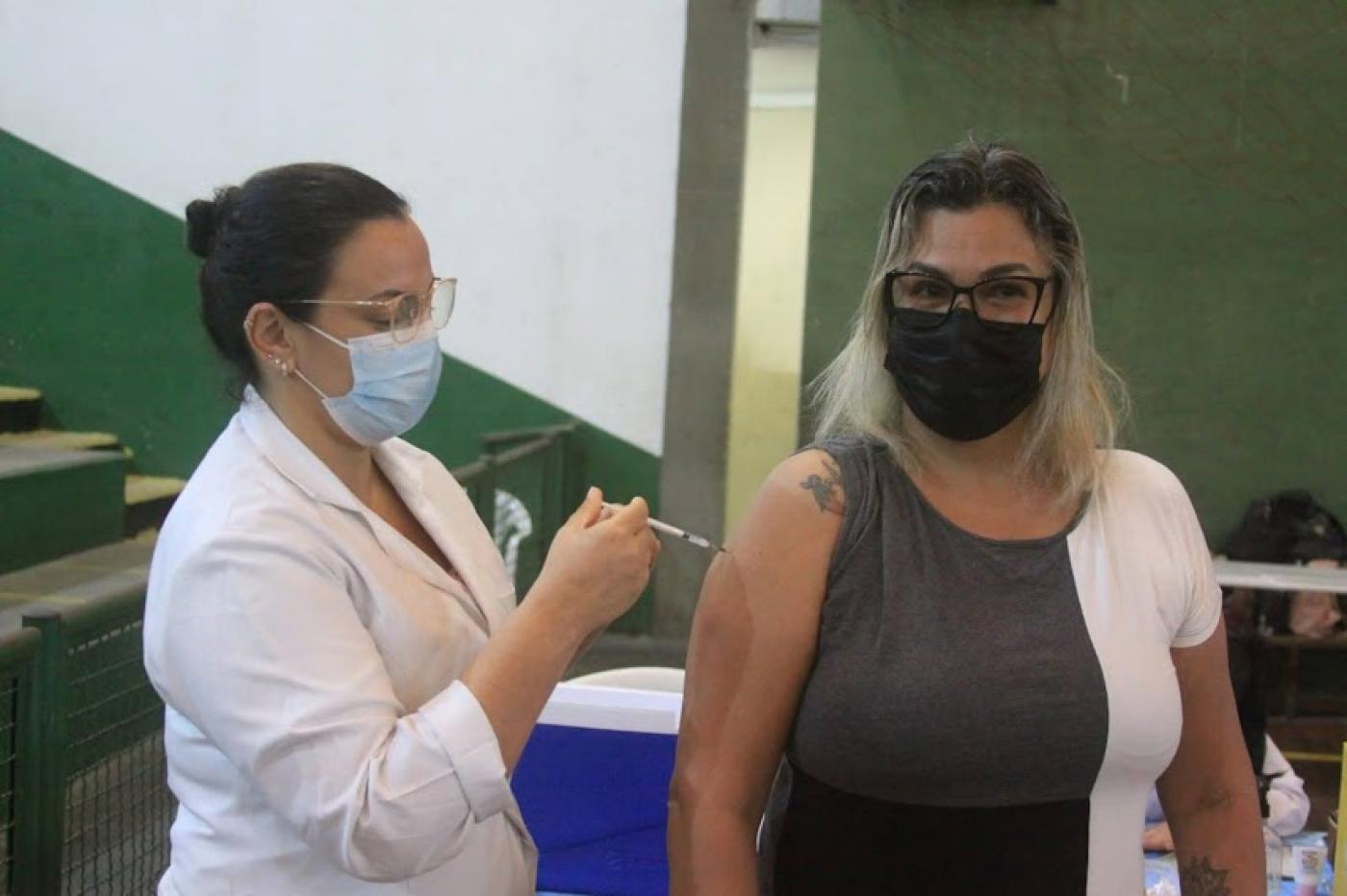 Mulher recebe aplicação de vacina no braço, feita por profissional de saúde. #pracegover
