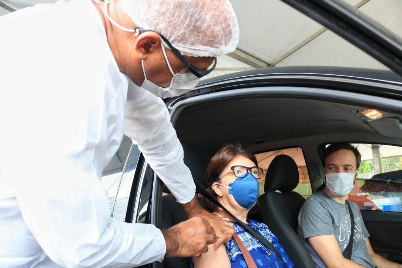 mulher é vacinada dentro do carro. Ela está no banco do passageiro ao lado do motorista. #paratodosverem