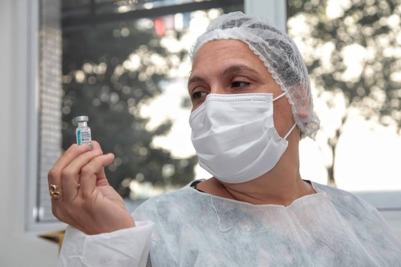 mulher usando avental, touca e máscara segura frasco com dose de vacina na mão direita. Ela olha o frasco.#paratodosverem