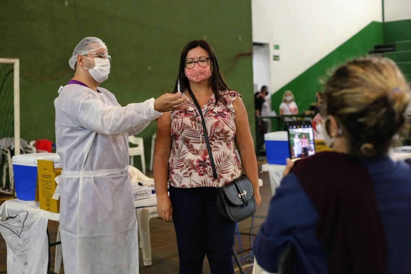 Enfermeiro mostra vacina para mulher e outra tira foto #paratodosverem