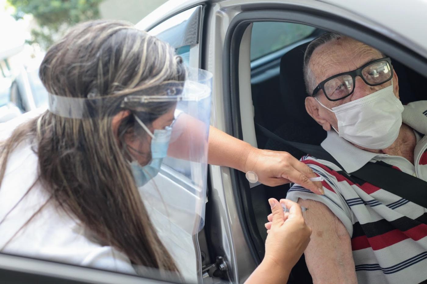 Idoso está dentro de carro sendo vacinado por uma mulher. #Paratodosverem