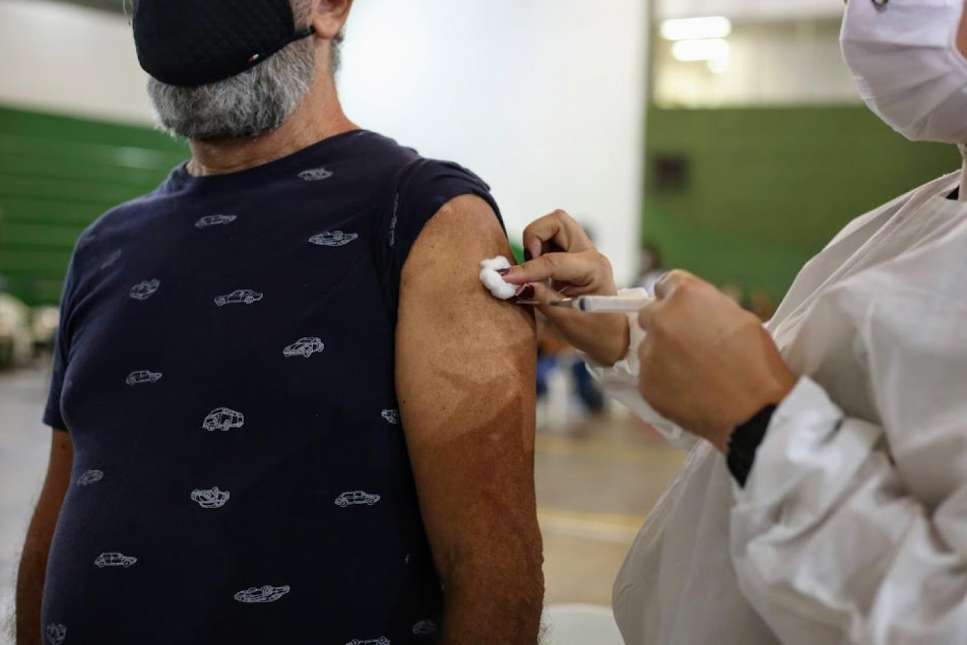 imagem em close de uma pessoa vacinando o braço de um homem, cuja camiseta está com a manga recolhida ao ombro. Ele usa máscara. #paratodosverem