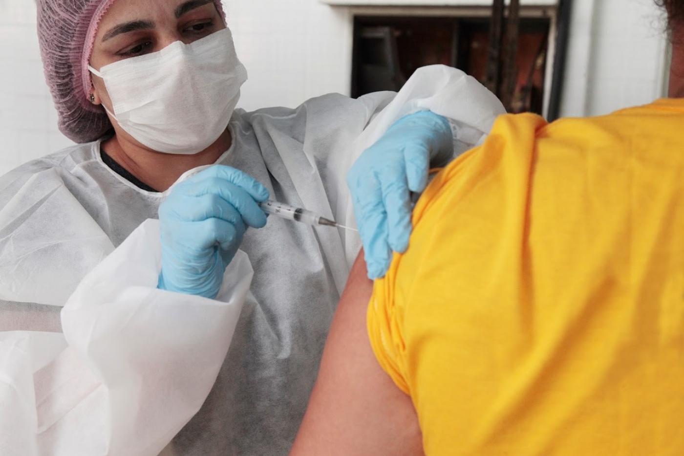 Mulher usando máscara, gorro, avental e luvas vacina uma pessoa que de costas, em close. #Paratodosverem