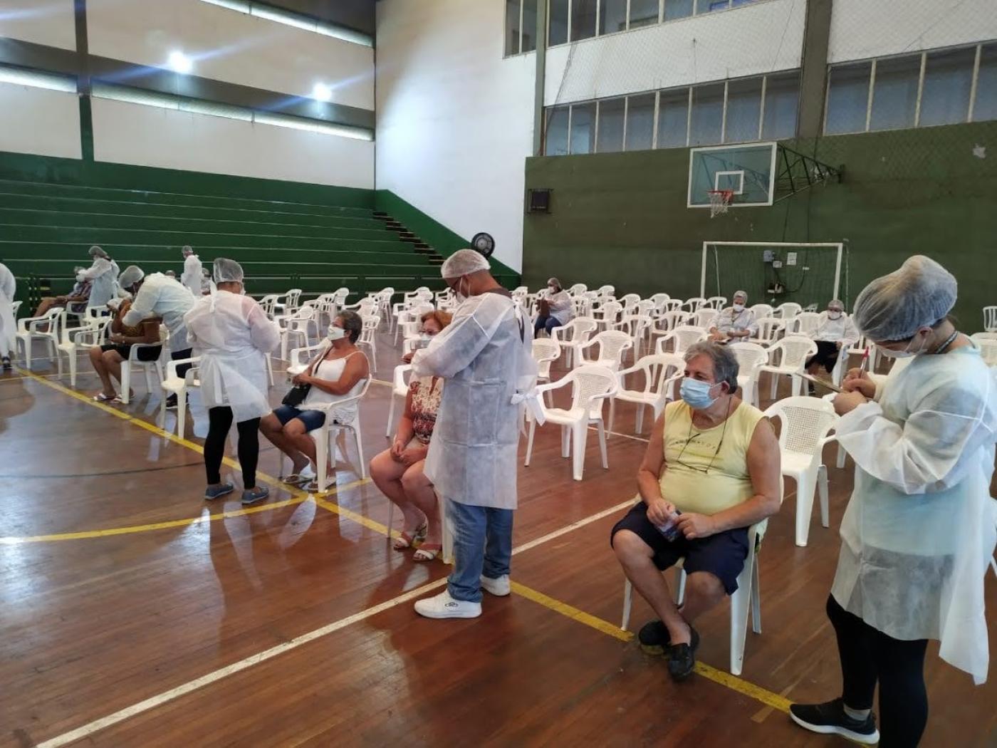 Ginásio de esportes com cadeiras enfileiradas. Na primeira filha há alguns idosos sentados aguardando a vacinação. Os aplicadores estão em pé. #paratodosverem