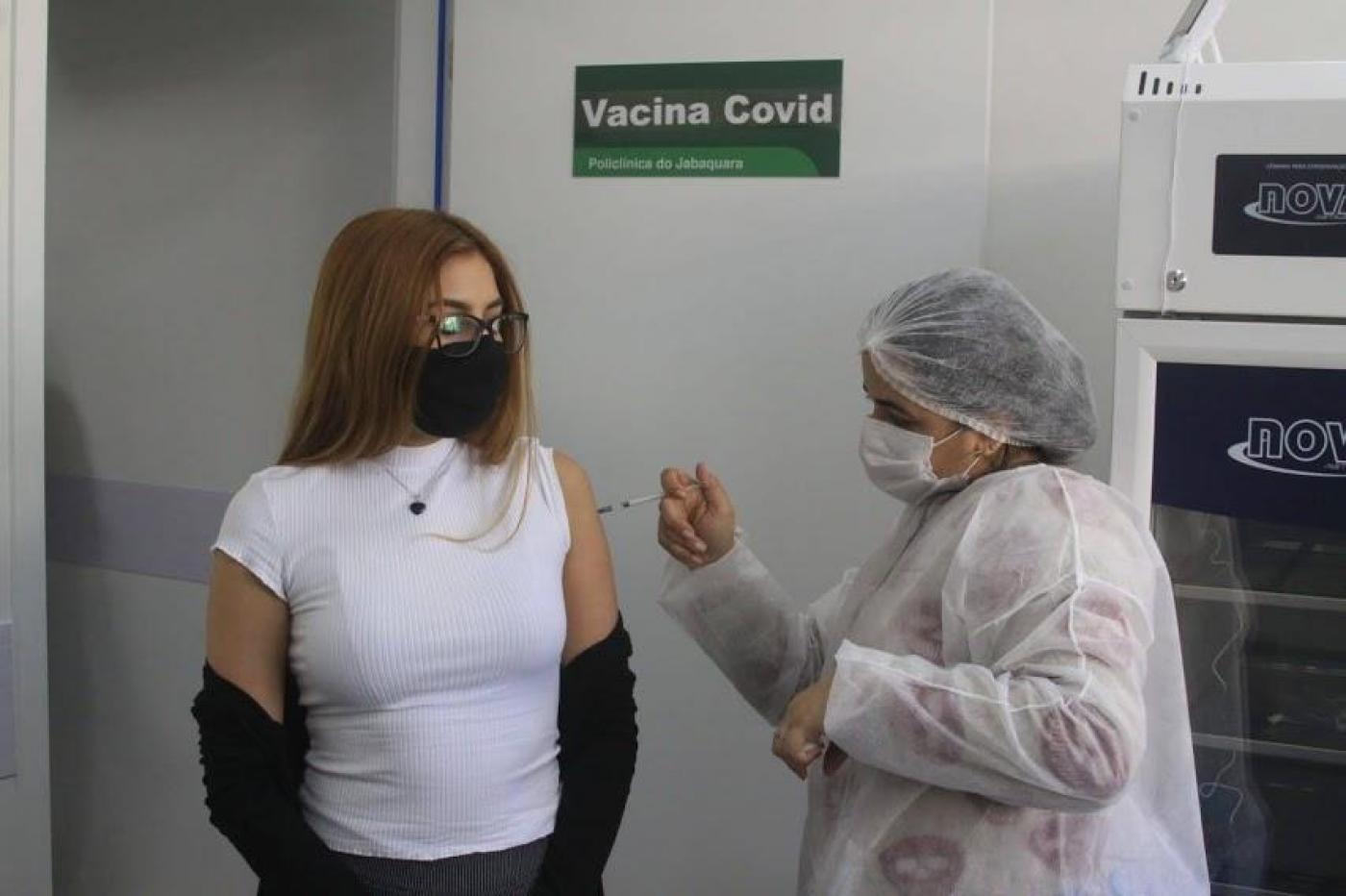 Jovem recebe aplicação de vacina por profissional de saúde paramentada. #pracegover