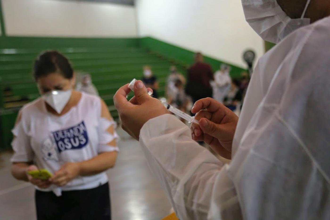 mulher prepara  vacina, em primeiro plano, e outra aguarda, segurando um celular. #paratodosverem