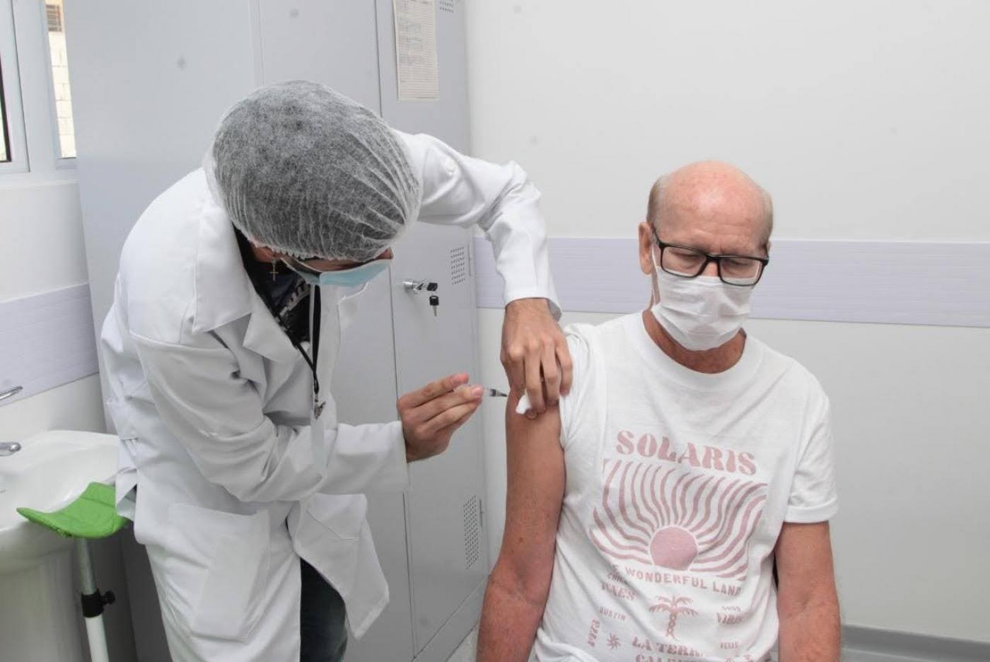 Homem está sentado em cadeira sendo vacinado por outro homem paramentado com avental, gorro e máscara. #Paratodosverem