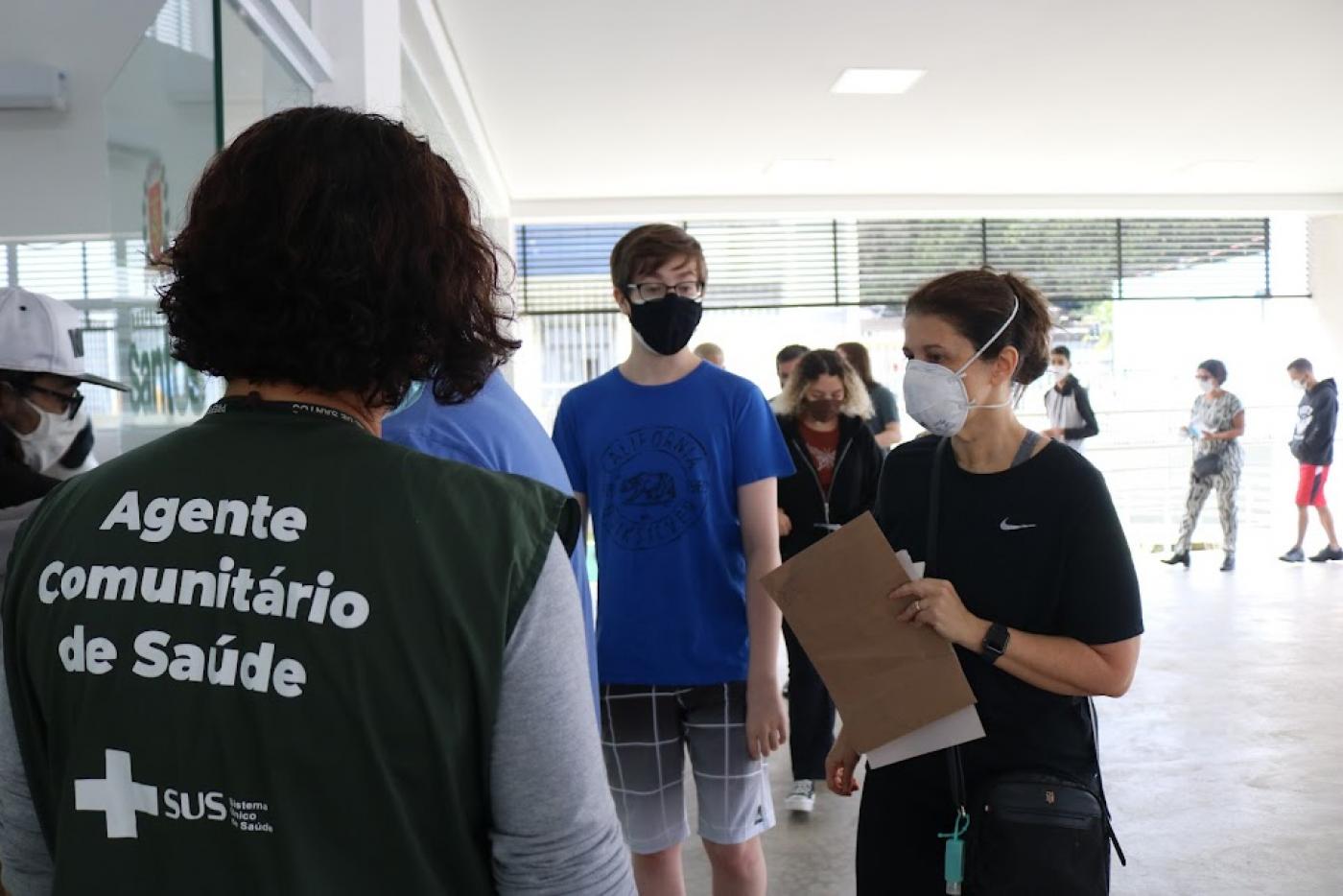 agente comunitário usando colete está em primeiro plano, de costas. Atrás, uma fila de adolescentes. #paratodosverem