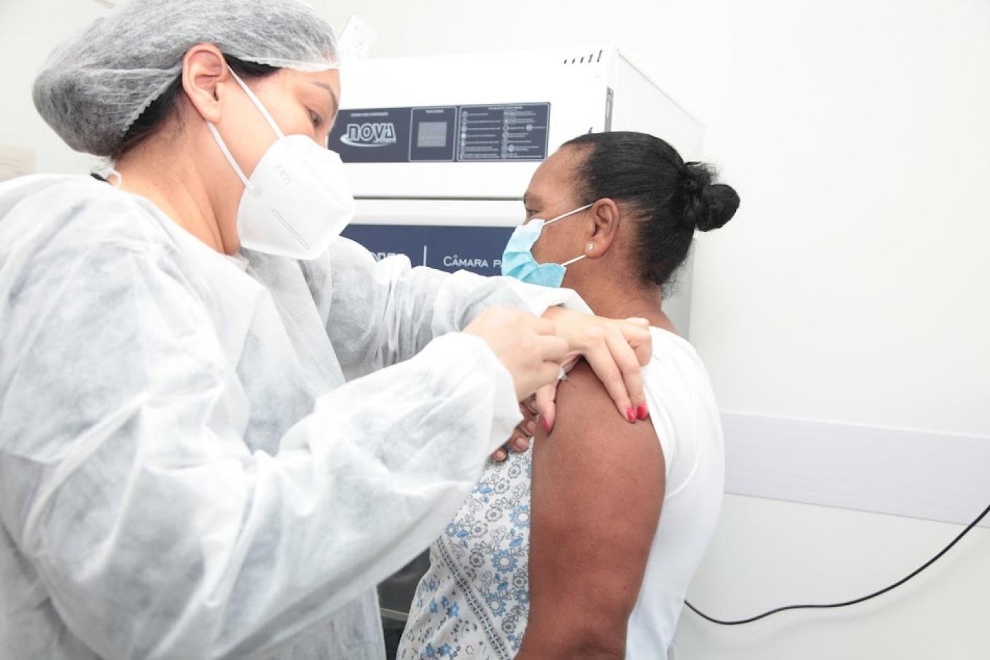 mulher usando avental touca e máscara injeta agulha em braço de outra mulher. #paratodosverem