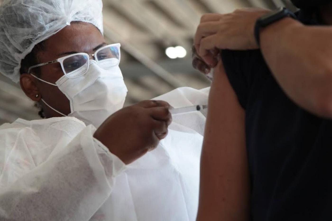 mulher usando máscara e gorro vacina o braço de uma pessoa que segura a manga da blusa. #paratodosverem