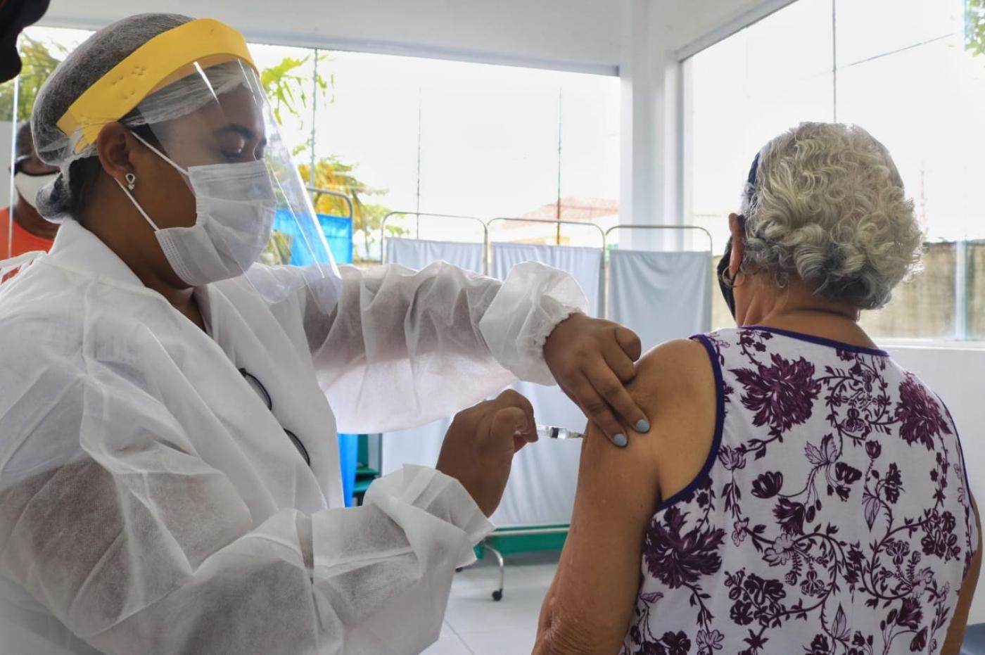 idosa está de costas sendo vacinada no braço esquerdo por uma mulher paramentada com avental, máscara e face shield. #Paratodosverem