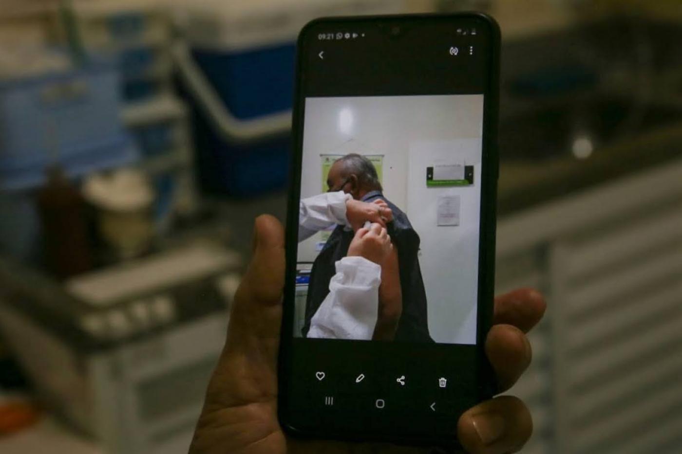 MÃO SEGURA SMARTPHONE QUE FOTOGRAFA HOMEM SENDO VACINADO. #paratodosverem