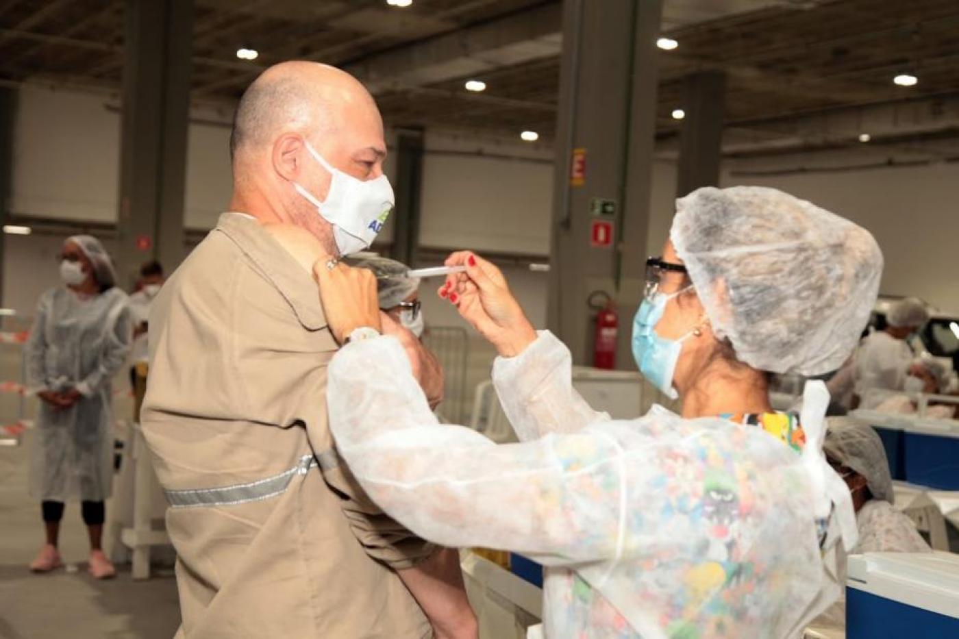 Homem recebe aplicação de vacina no braço por profissional de saúde. #pracegover