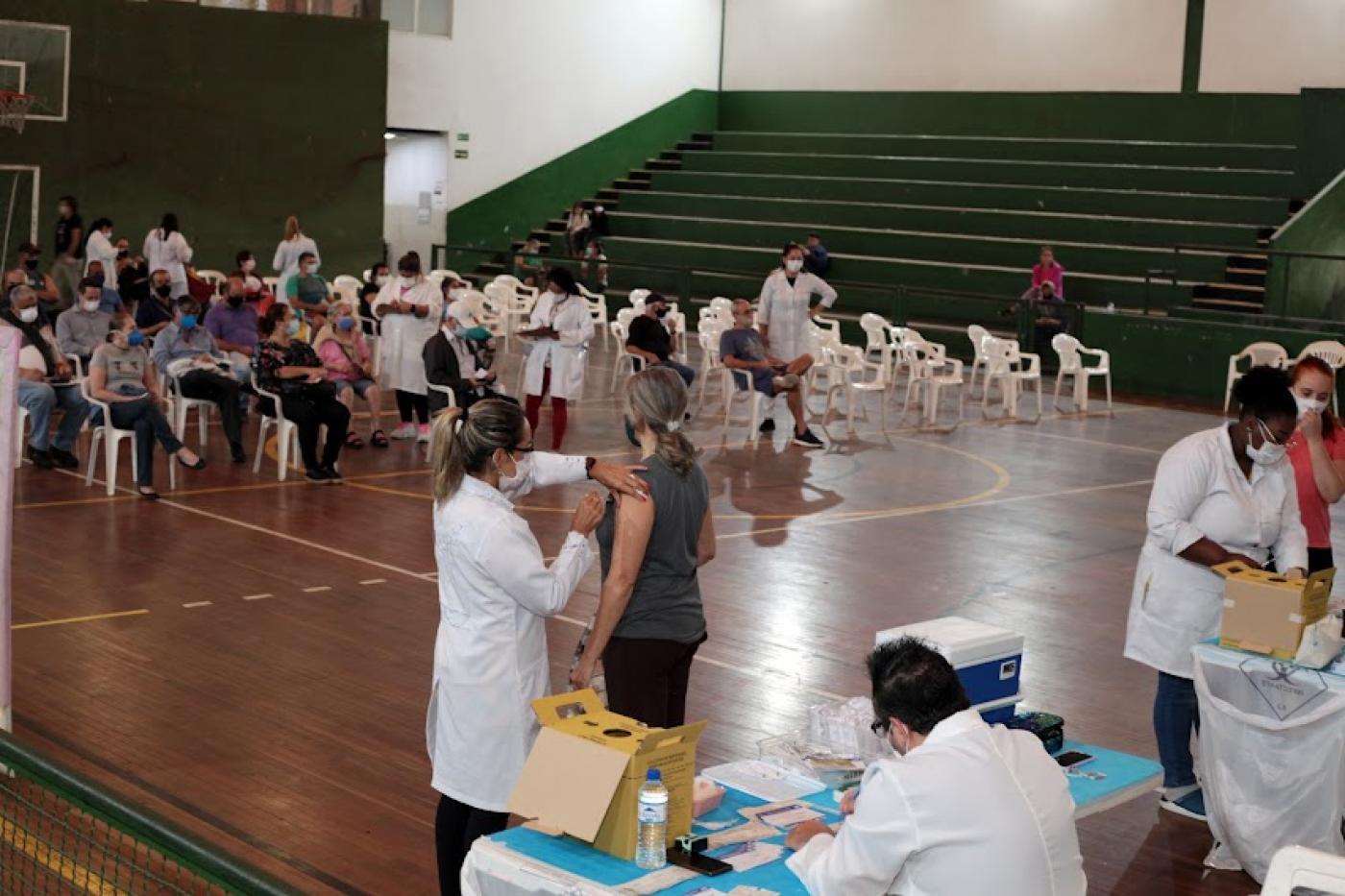 público está sentado em quadra de ginásio aguardando atendimento. Em primeiro plano, mulheres aplicam vaciina em pessoas em pé. #paratodosverem