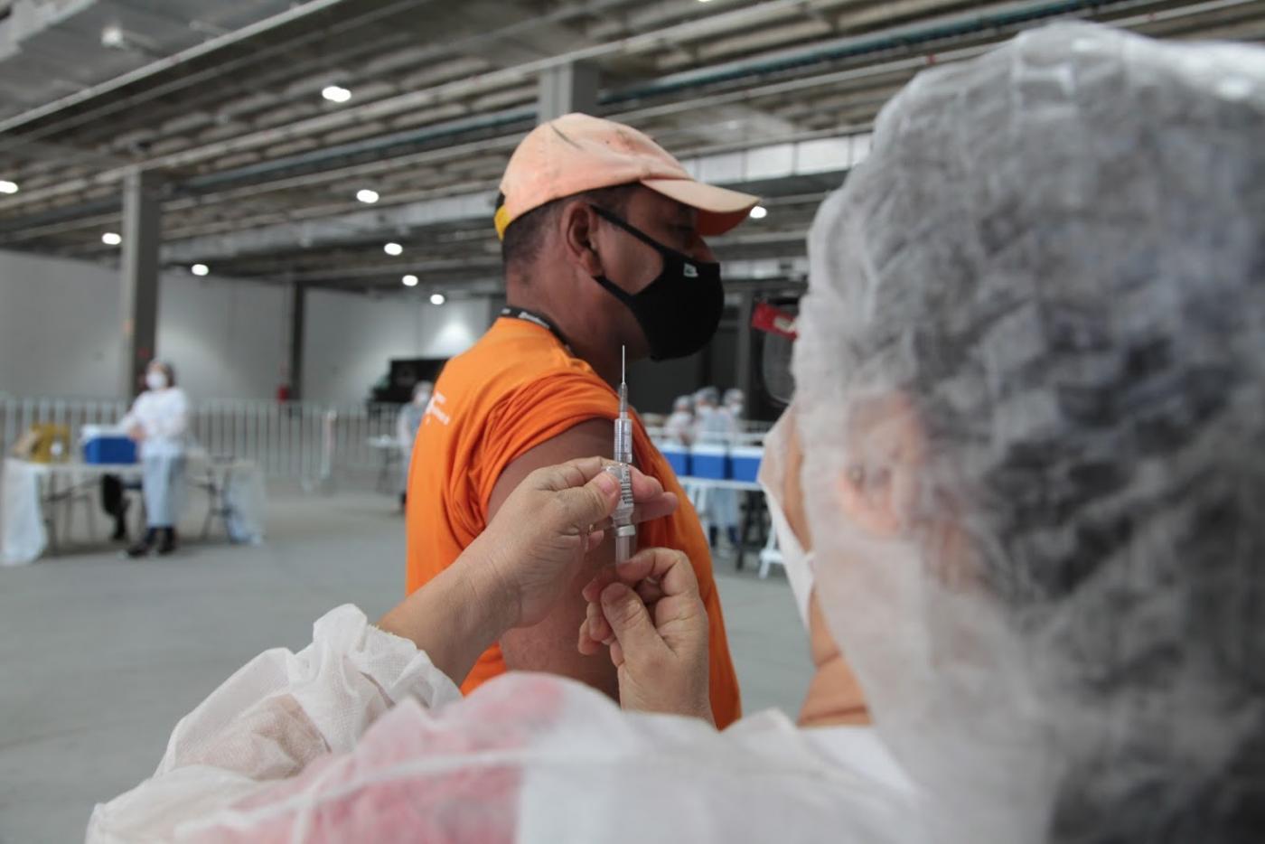 mulher segura seringa com agulha, se preparando para vacina homem que está com a manga da camisa arregaçada. #paratodosverem