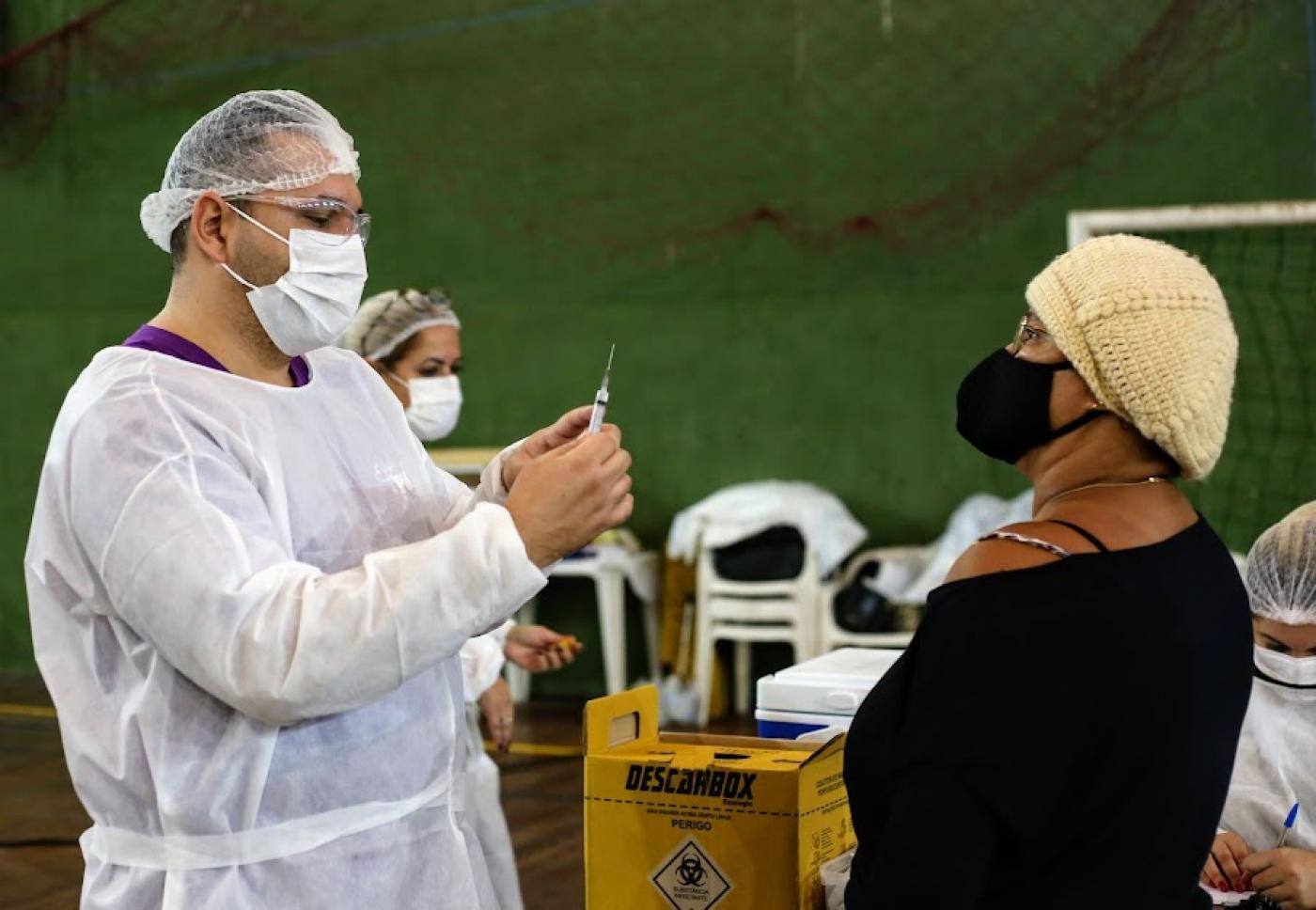 homem segura seringa de vacina diante de uma mulher. #paratodosverem