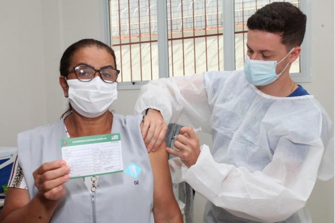 Trabalhadora da saúde mostra carteira de vacinação enquanto recebe vacina no braço por profissional paramentado. #pracegover