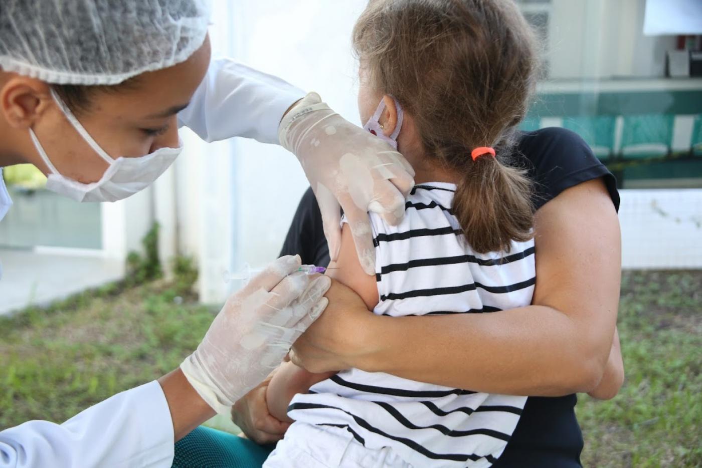 Criança está de costas, no colo, enquanto é vacinada. #Paratodosverem