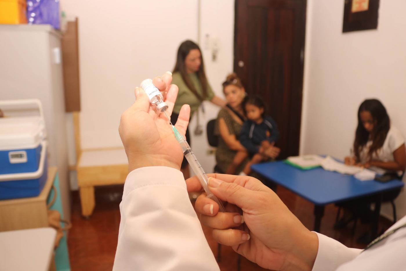 mão segura seringa em primeiro plano. Mulher e criança ao fundo. #paratodosverem