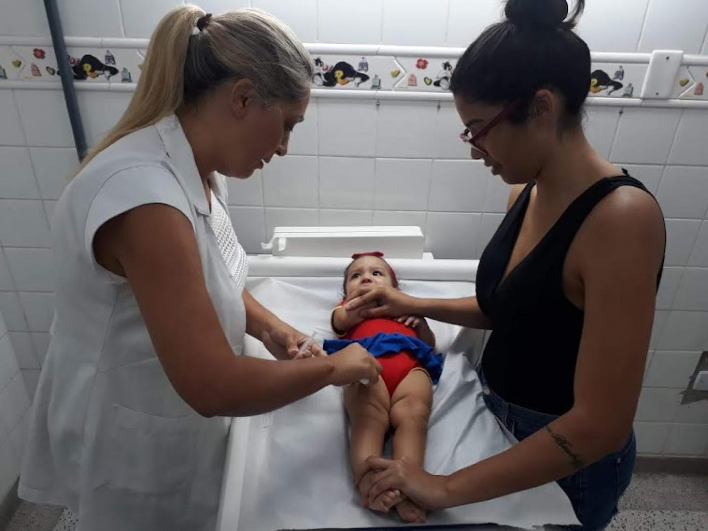 bebê está deitada em maca. Mulher aplica vacina e mãe segura a menina. #paratodosverem