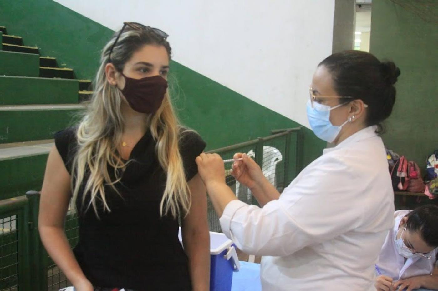 Mulher é vacina por profissional de saúde em ginásio poliesportivo. #pracegover