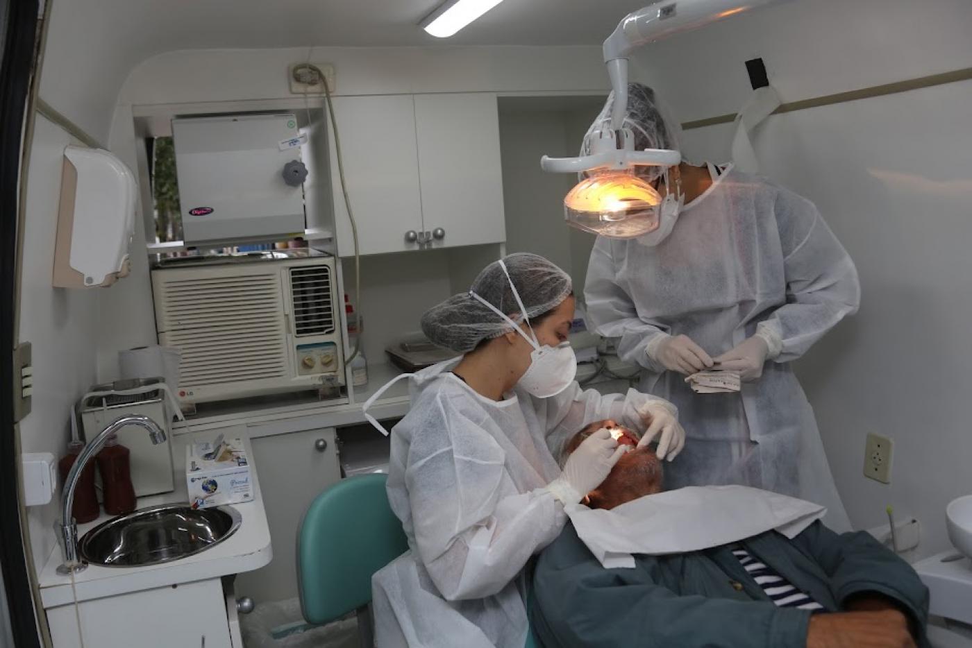 van odontológica equipada. Mulher examina um homem que está na cadeira. Outro homem, auxiliar do serviço, está em pé. #paratodosverem