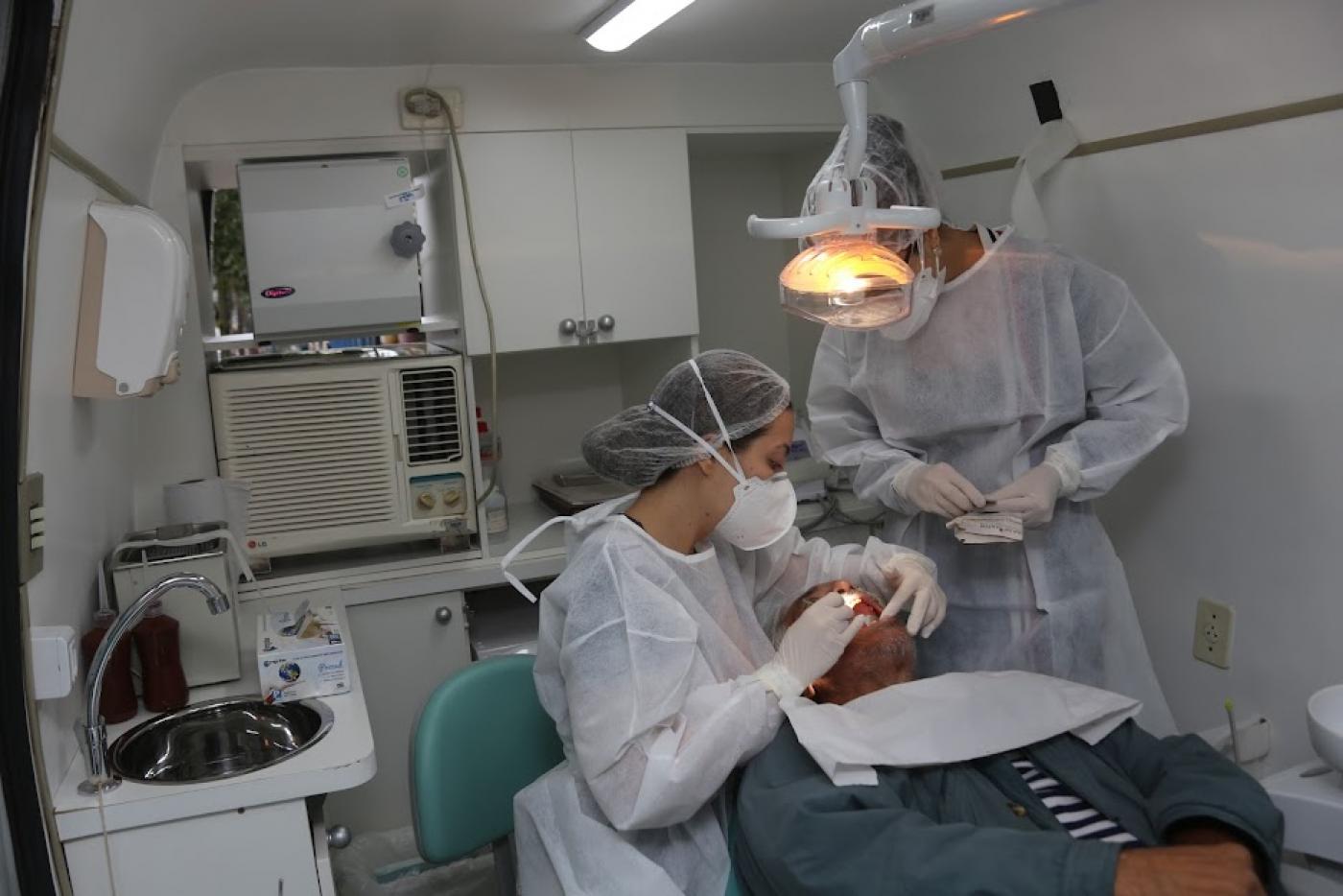 van odontológica equipada. Mulher examina um homem que está na cadeira. Outro homem, auxiliar do serviço, está em pé. #paratodosverem