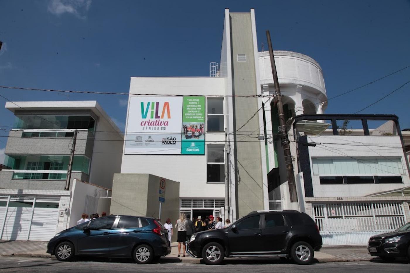 Fachada de vila criativa com placa no alto e carros estacionados na porta. #paratodosverem